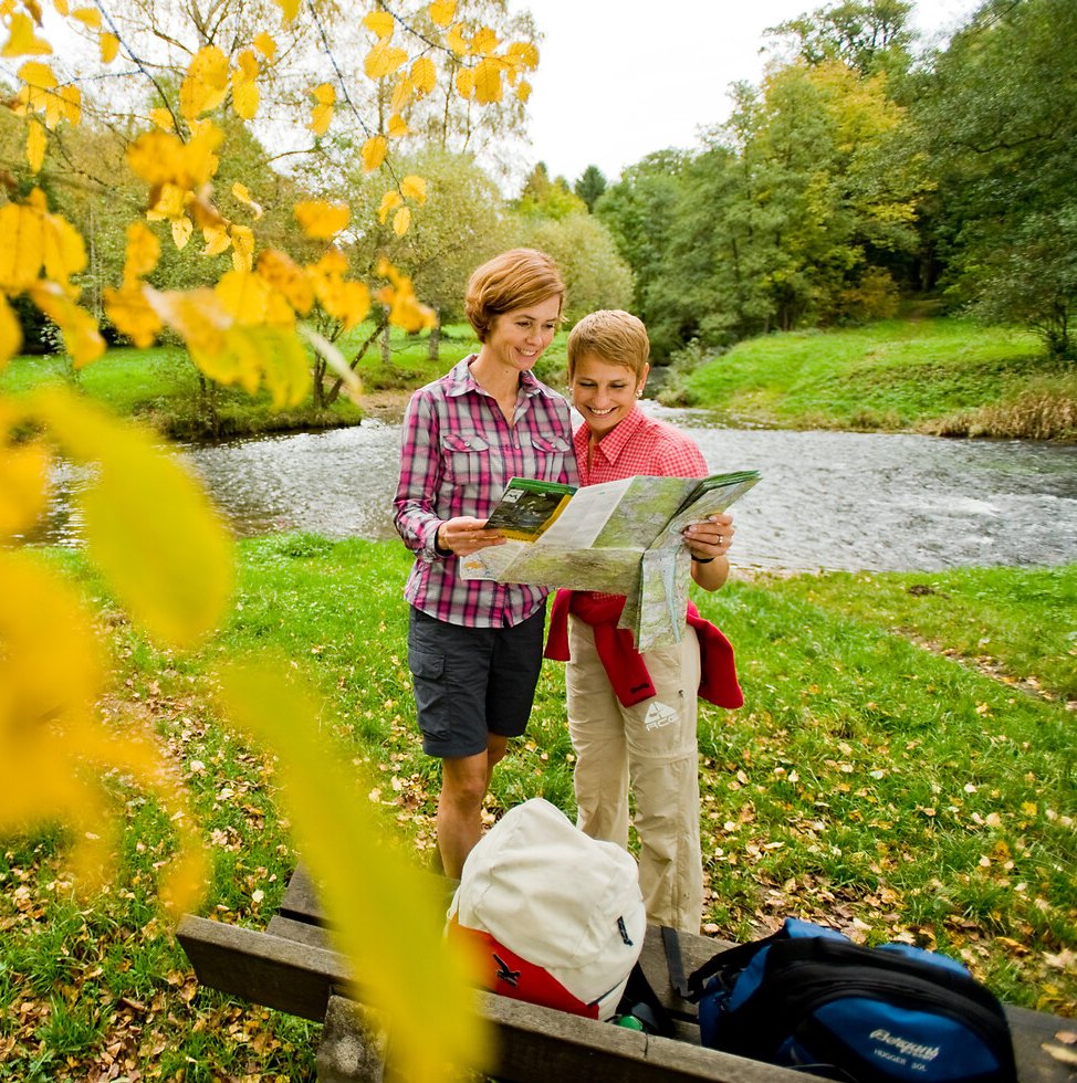 Zwei Wanderer rasten im Herbst an einer Bank am Fluss und schauen in die Wanderkarte