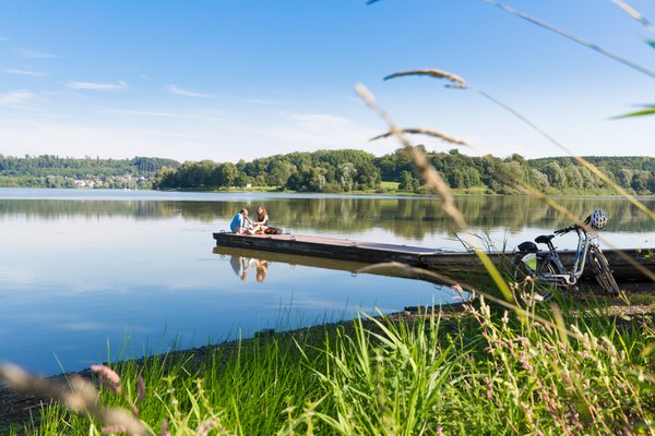 Zwei Personen sitzen auf einem Holzsteg am See, Fahrrad daneben