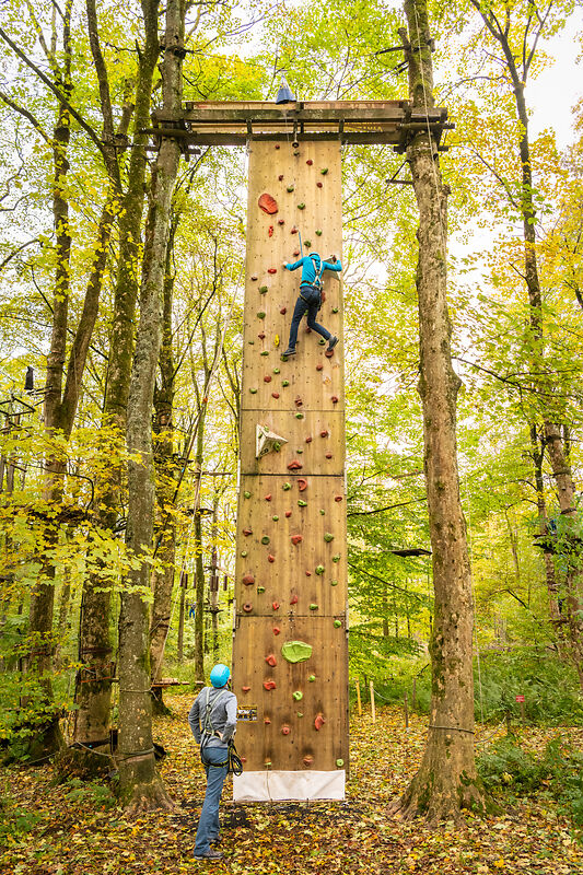 Person klettert an einer hohen Kletterwand in einer herbstlichen Landschaft